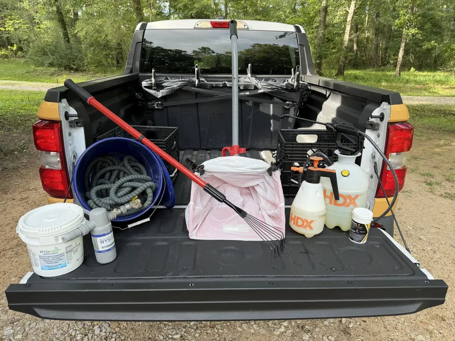 Open truck bed with a Wysiwash sanitizer jug, two pump sprayers, a long-handled rake, a lined waste bin, and a coiled hose