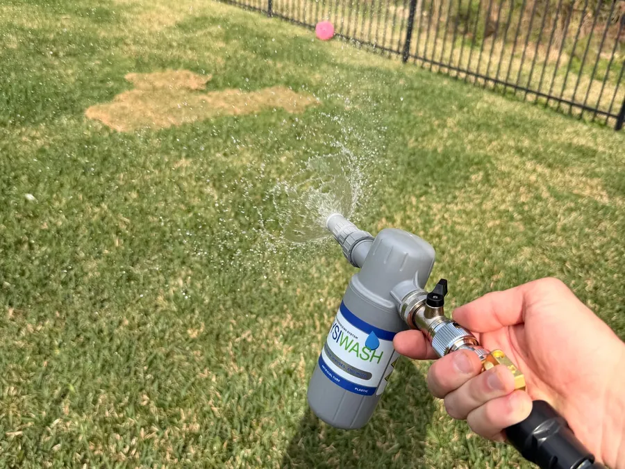 A technician's hand spraying a fenced backyard lawn with a Wysiwash hose-end sanitizer applicator